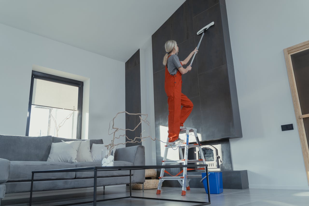 services-03 A woman stands on a ladder in a modern living room, cleaning the high wall with a mop.